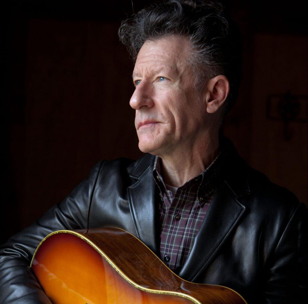 man with dark hair wearing dark leather jacket sitting with guitar looking pensively out with a side profile