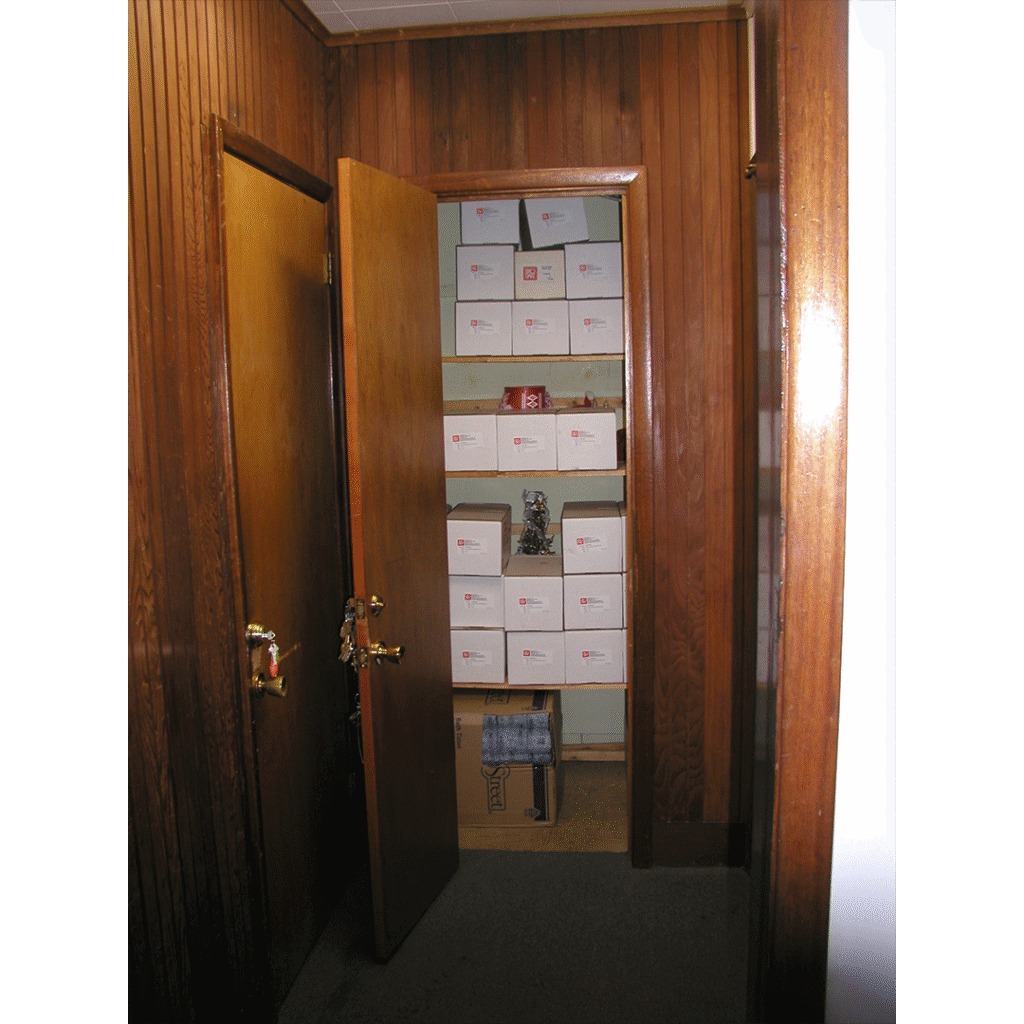 A small storage room inside the old Bruce Springsteen Archives, viewed through a partially open wooden door. The shelves are lined with white archival boxes labeled with red logos, and the walls and door frames are covered in dark wood paneling.