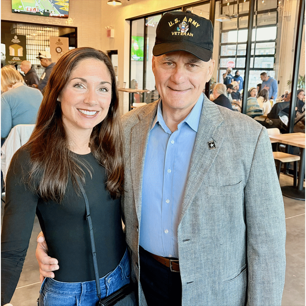 A woman and a man stand smiling in a busy cafe. The man wears a "U.S. Army Veteran" cap and a gray jacket. The scene is warm and social.