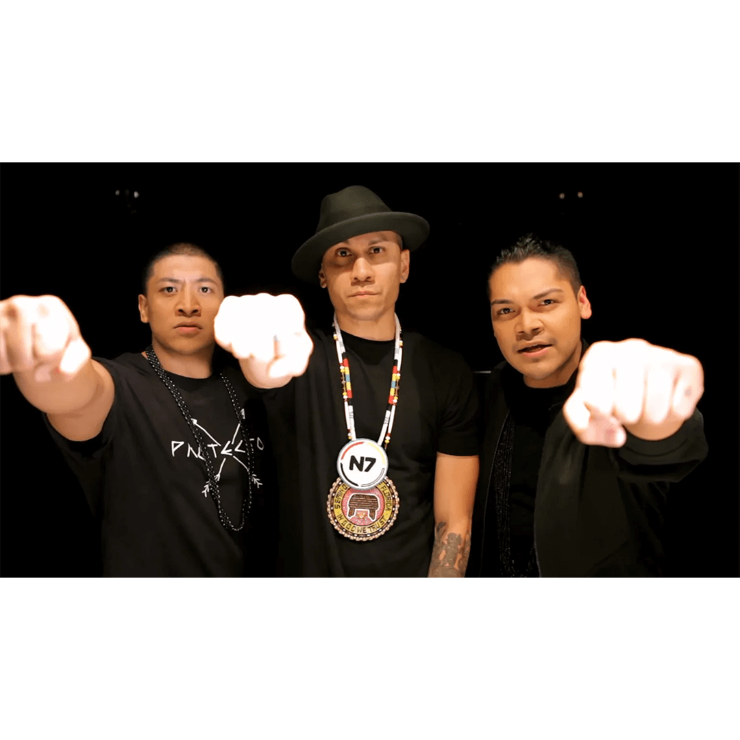 Three individuals facing forward, fist-bumping the camera. The central figure wears a fedora and beaded necklace. The image exudes confidence and unity.