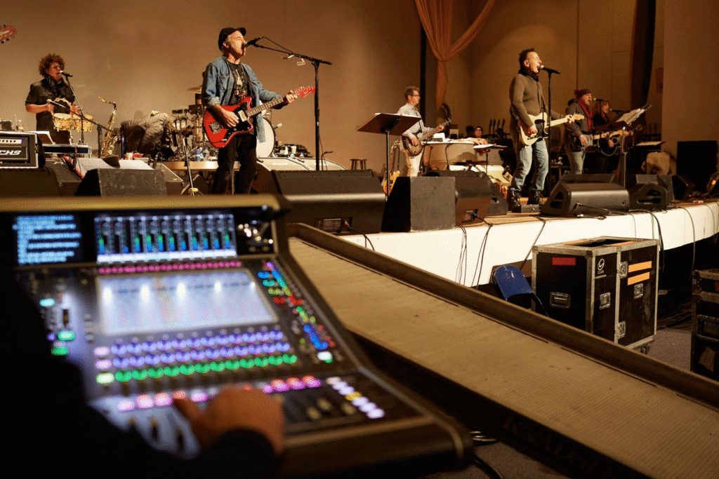 A band rehearses on stage, with musicians playing guitars and singing into microphones. In the foreground, a person adjusts a colorful audio mixing board.