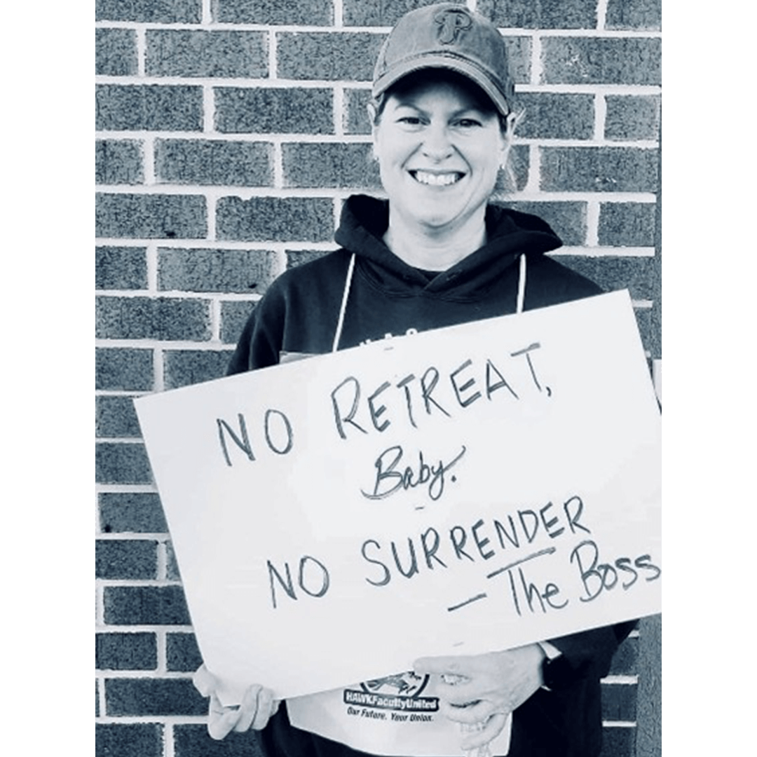 Smiling person in a cap holds a sign reading "No Retreat, Baby. No Surrender - The Boss" against a brick wall. The mood is determined and encouraging.