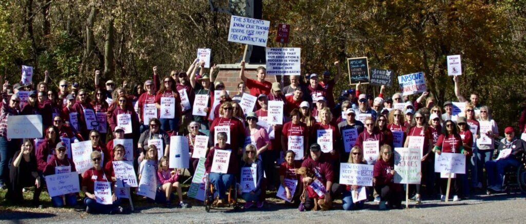 A group of people wearing maroon shirts and holding signs gather outdoors for a protest. Signs advocate for fair contracts and education support. The mood is determined and unified.