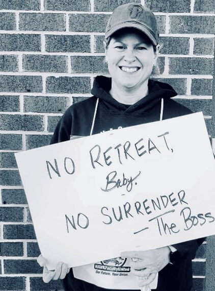 Smiling person in a cap holds a sign reading "No Retreat, Baby. No Surrender - The Boss" against a brick wall. The mood is determined and encouraging.