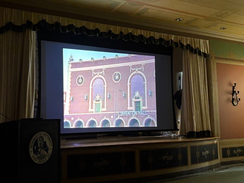 A large screen in a dimly lit room displays an image of a building labeled "Convention Hall" with arched windows. A podium with a university seal is on the left. The setting is formal.