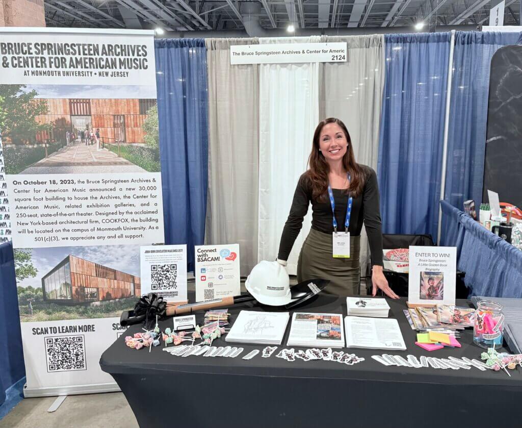 A woman stands smiling behind a booth for the Bruce Springsteen Archives & Center for American Music. The table displays brochures, QR codes, and promotional items, including a branded hard hat and small toys. The backdrop features informational posters about the center. The setting conveys an inviting and informative atmosphere at a convention.