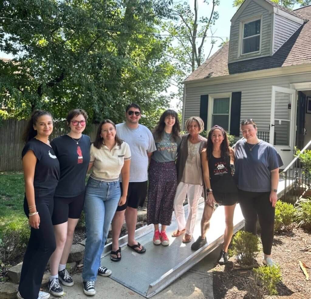 A group of eight people stands together outside a house on a sunny day, smiling warmly. Trees and the house create a welcoming, friendly atmosphere.