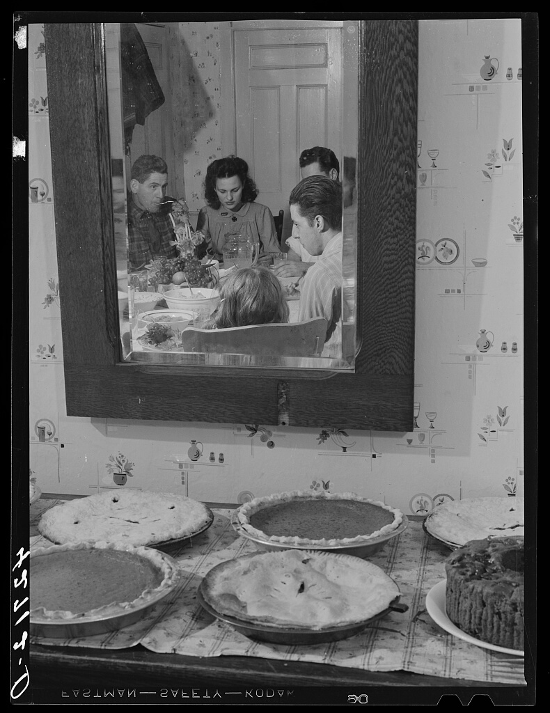 Reflection of a family dining around a table, seen in a wall mirror. In the foreground, a table displays several pies and a cake, evoking a cozy holiday meal.