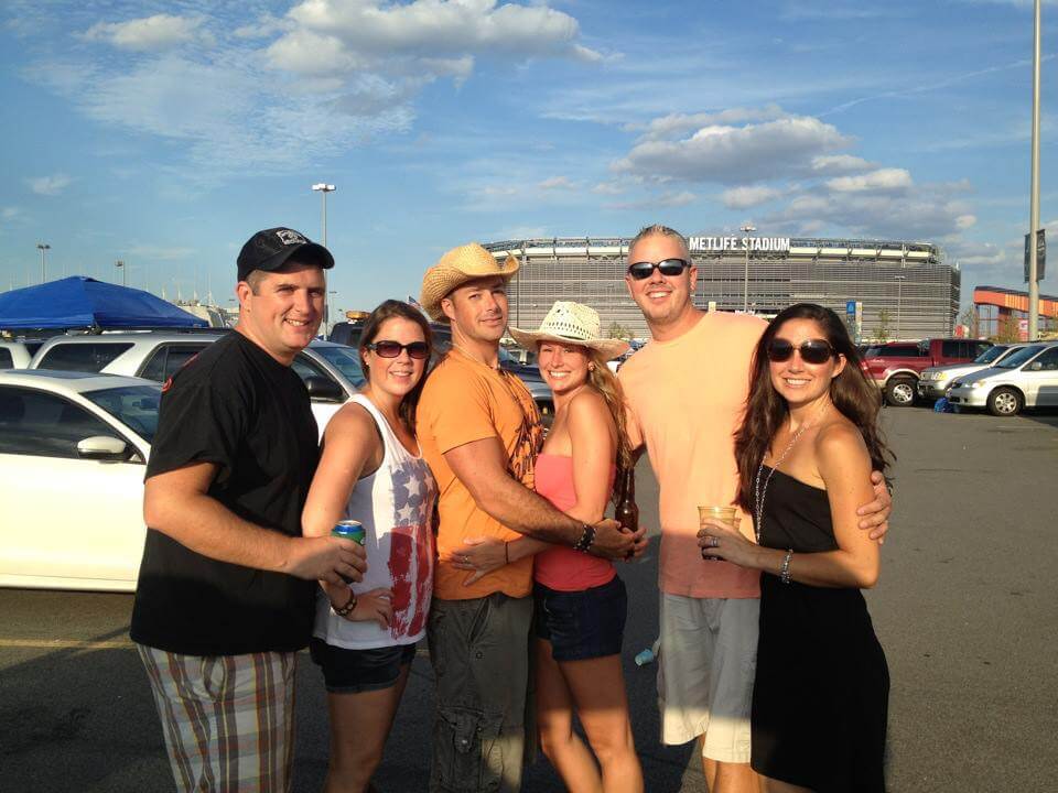 A group of six people smiling in a parking lot with MetLife Stadium in the background. They wear casual summer clothes, some in sunglasses and hats, conveying a cheerful atmosphere.