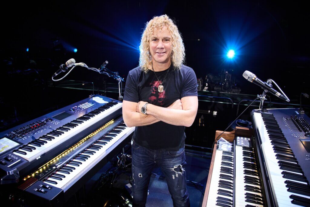 A person with curly blond hair smiles while standing between two keyboards on a dark stage. Bright lights shine in the background, creating a vibrant atmosphere.
