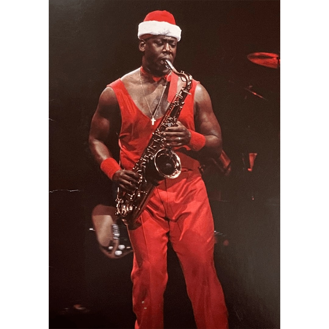 A man in a Santa hat and red outfit plays a saxophone on stage, conveying a festive and energetic mood. The spotlight highlights his concentration.
