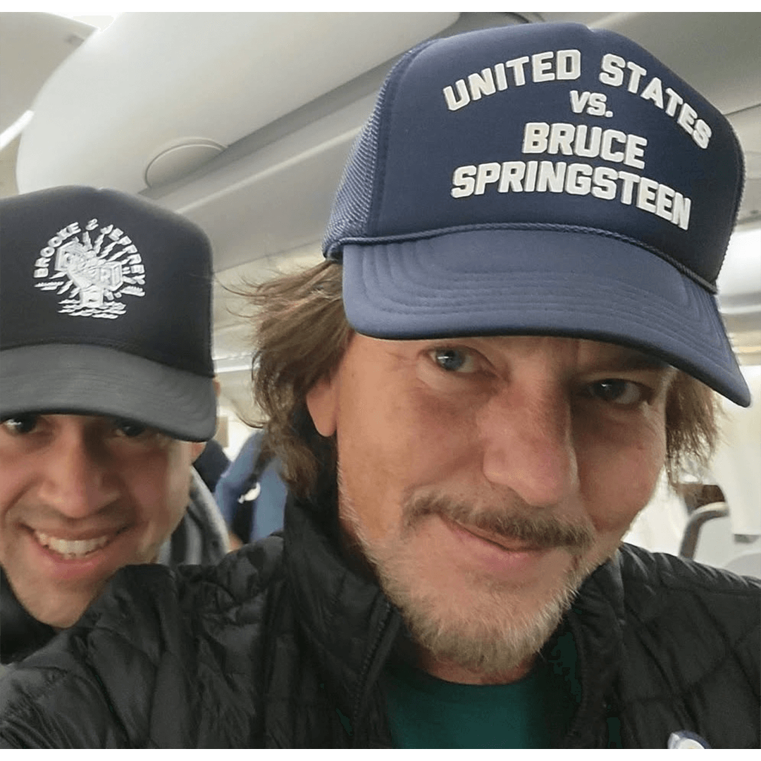 Two men smiling inside an airplane. The man in front wears a cap reading "United States vs. Bruce Springsteen," creating a casual and humorous tone.