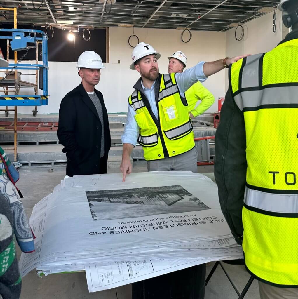 A group of construction workers and a man in a suit discuss architectural blueprints on-site. The man pointing at the plans wears a white hard hat and yellow safety vest, highlighting collaboration and focus in a partially constructed room.