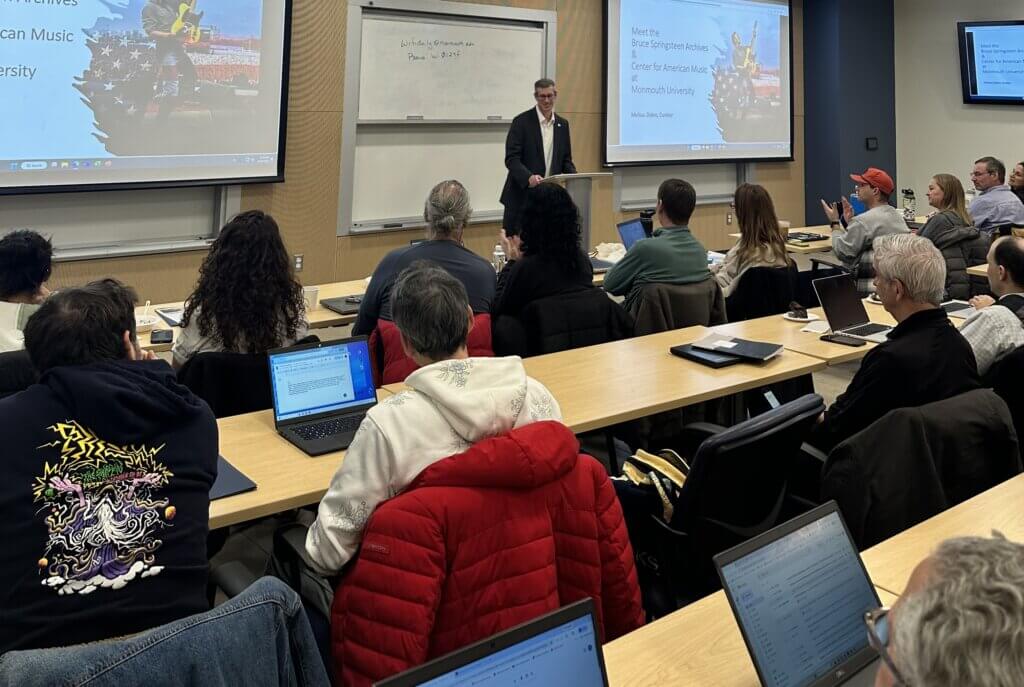 A classroom with people sitting at desks using laptops while a speaker stands at the front near a presentation screen. The atmosphere is focused and engaged.
