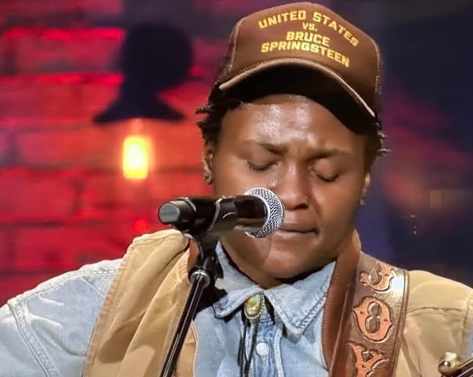 Singer in denim shirt and tan jacket plays guitar on stage, eyes closed, wearing a hat reading "United States vs. Bruce Springsteen," against a warm, glowing backdrop.