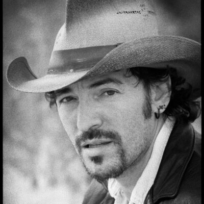 Bruce Springsteen with facial hair, earring and cowboy hat suede jacket and white collared shirt