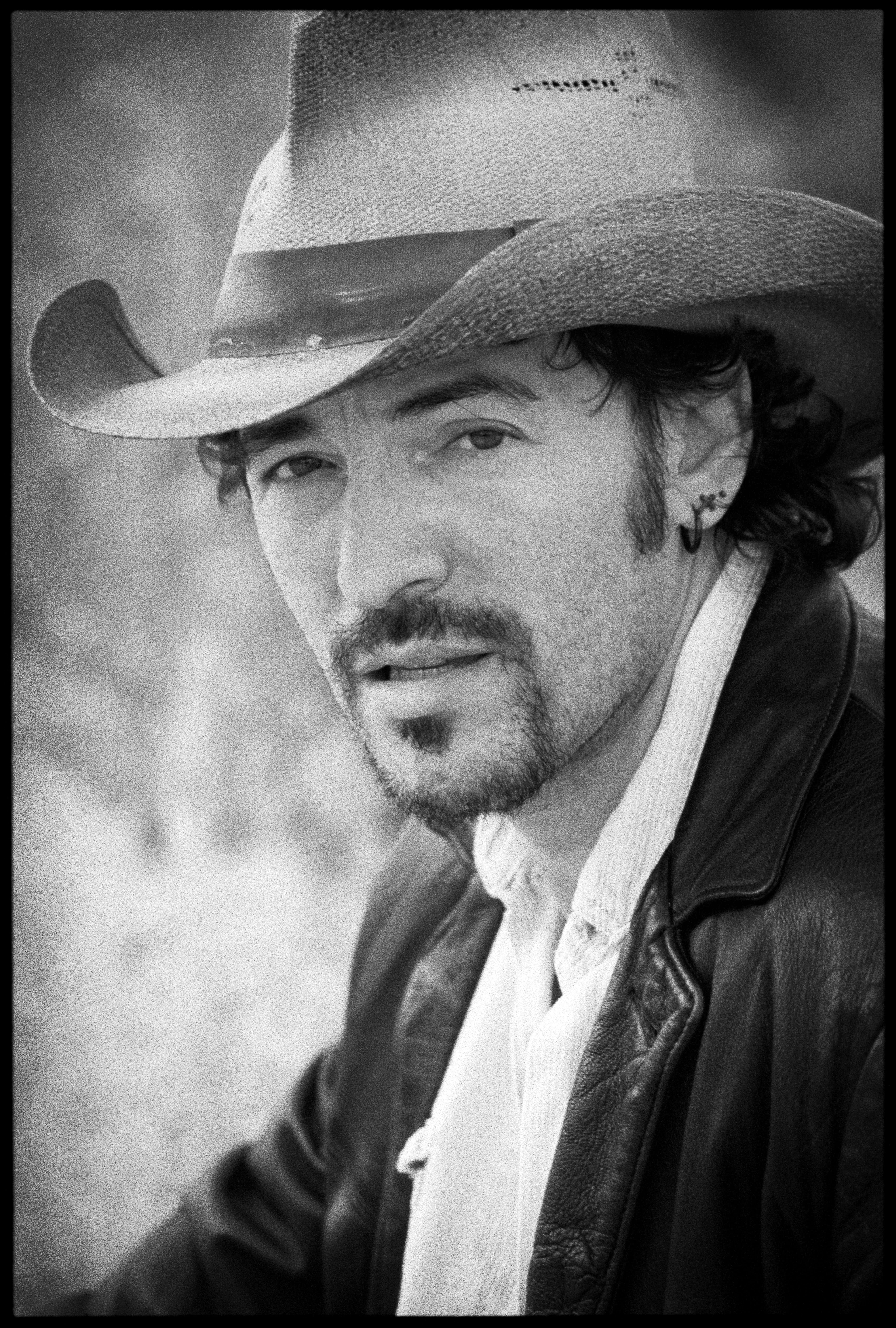 Bruce Springsteen with facial hair, earring and cowboy hat suede jacket and white collared shirt