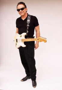 Man wearing black shirt and black pants with a guitar strapped around his body casually and wearing dark sunglasses