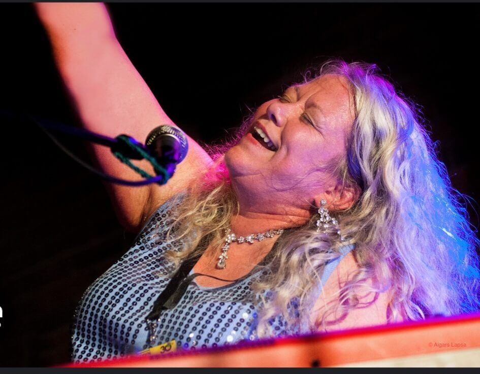 Woman with gray hair eyes closed raising arm up in the air, smiling and singing into microphone sitting down