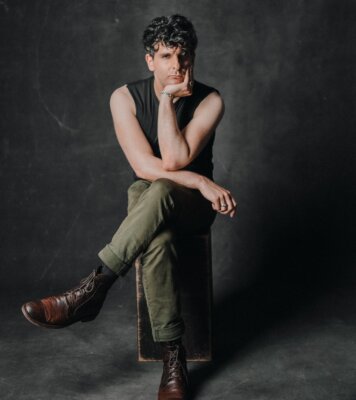 man with curly hair sitting crossed legged on a stool with his chin in his hand looking forward