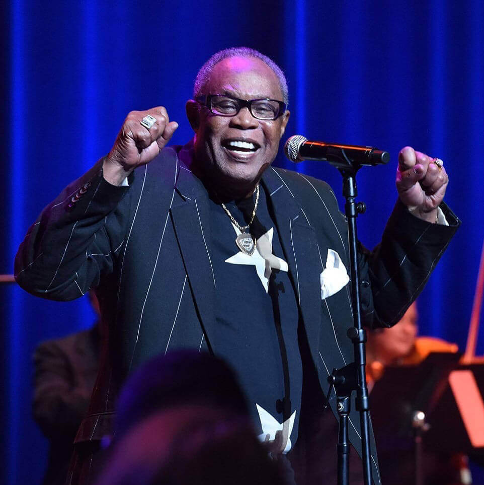 black man smiling with fists in the air at microphone wearing silver stars on jacket and eyeglasses