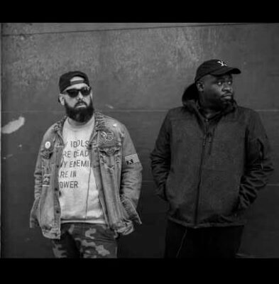 2 men leaning against a wall with hands in pockets wearing caps on heads. one is wearing dark glasses. both are wearing jackets. one man has a beard.