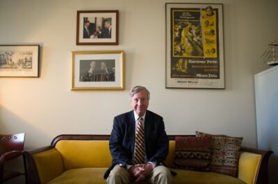 gray haired man sitting on yellow sofa wearing suit and tie with photos and painting on wall behind him